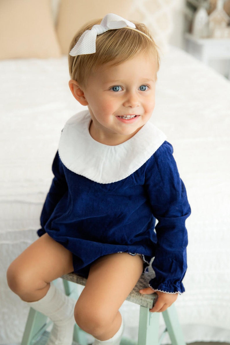 May include: A young child wearing a navy blue dress with a white collar and white ruffled sleeves. The dress has a white trim around the bottom. The child is wearing a white bow headband and has blonde hair. The child is sitting on a light blue stool.