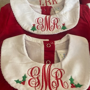 May include: Red corduroy dress with a white Peter Pan collar and a red embroidered monogram with a holly design. The monogram is "SMR".