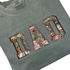 May include: Green camouflage print "DAD" graphic on a green t-shirt.