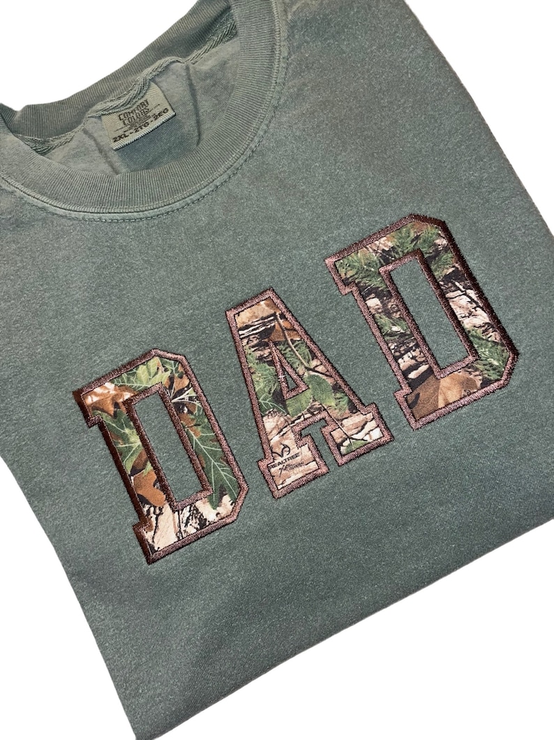 May include: Green camouflage print "DAD" graphic on a green t-shirt.