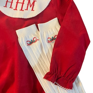 May include: Red corduroy baby onesie with a white Peter Pan collar and the monogram "HHM" embroidered on the collar. The onesie is paired with white ribbed knit socks with a green holly and red bow design.