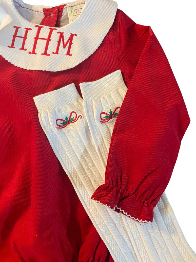May include: Red corduroy baby onesie with a white Peter Pan collar and the monogram "HHM" embroidered on the collar. The onesie is paired with white ribbed knit socks with a green holly and red bow design.