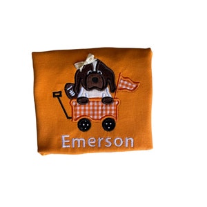 Puede incluir: Camiseta naranja con un dise&ntilde;o bordado que presenta un perro marr&oacute;n y blanco en un vag&oacute;n. El vag&oacute;n tiene un patr&oacute;n de cuadros y una peque&ntilde;a bandera. La palabra "Emerson" est&aacute; bordada en blanco debajo.