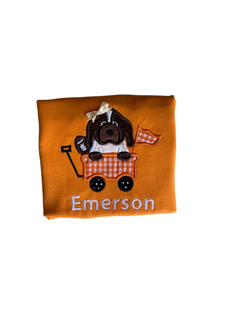 Puede incluir: Camiseta naranja con un dise&ntilde;o bordado que presenta un perro marr&oacute;n y blanco en un vag&oacute;n. El vag&oacute;n tiene un patr&oacute;n de cuadros y una peque&ntilde;a bandera. La palabra "Emerson" est&aacute; bordada en blanco debajo.