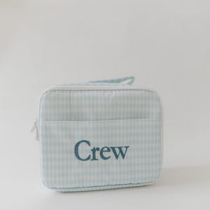 May include: A rectangular zippered pouch featuring a light blue and white gingham pattern. The front of the pouch includes an open pocket with the word Crew embroidered in dark blue thread. The pouch has white piping along the edges and a handle.