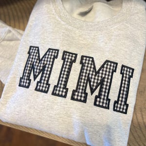 Puede incluir: Sudadera gris claro con la palabra "MIMI" bordada en el pecho en tela de cuadros blancos y negros. La sudadera está doblada sobre una superficie de madera.