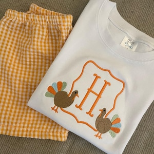 May include: A white t-shirt with a monogrammed "H" in an orange frame with two embroidered turkeys on either side. The shirt is paired with orange and white gingham shorts.