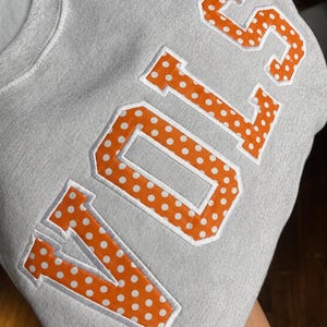 Peut inclure: Un sweat-shirt gris clair à col rond arborant le mot VOLS en grandes lettres de style bloc. Les lettres sont orange avec un motif à pois blancs et sont soulignées par une broderie blanche sur le tissu gris du vêtement.