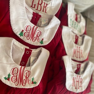 May include: Five red Christmas dresses with white collars and embroidered monograms. Each dress has a green holly sprig with red berries embroidered on the collar. The monograms are LBK, SMR, TEZ, MHR, and NHR.