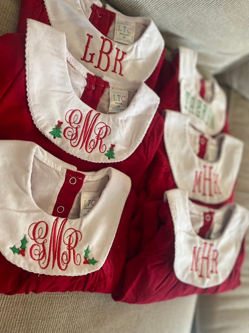 May include: Five red Christmas dresses with white collars and embroidered monograms. Each dress has a green holly sprig with red berries embroidered on the collar. The monograms are LBK, SMR, TEZ, MHR, and NHR.