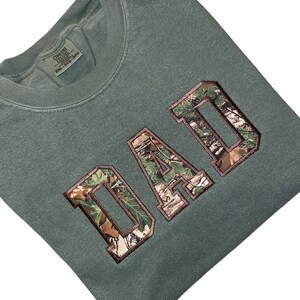 May include: A green t-shirt with the word "DAD" in large, brown letters with a camouflage pattern. The letters are outlined in brown.