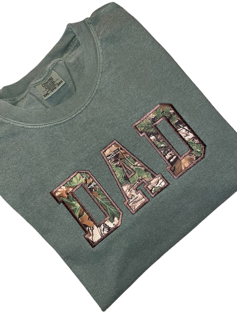 May include: A green t-shirt with the word "DAD" in large, brown letters with a camouflage pattern. The letters are outlined in brown.