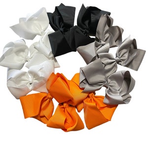 Puede incluir: Una colecci&oacute;n de lazos grandes para el cabello, dispuestos en forma circular. Los lazos son de color blanco, negro, gris y naranja. Cada lazo est&aacute; hecho de un material liso, similar al sat&eacute;n, y atado en forma de lazo cl&aacute;sico.