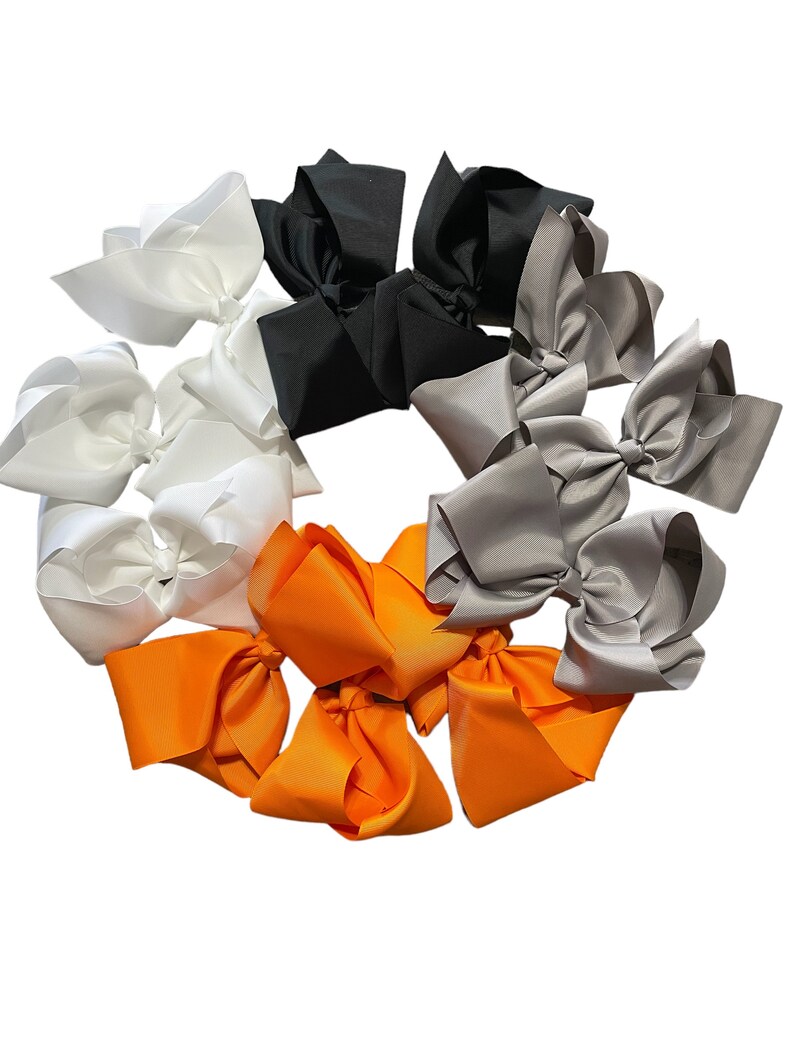 Puede incluir: Una colecci&oacute;n de lazos grandes para el cabello, dispuestos en forma circular. Los lazos son de color blanco, negro, gris y naranja. Cada lazo est&aacute; hecho de un material liso, similar al sat&eacute;n, y atado en forma de lazo cl&aacute;sico.