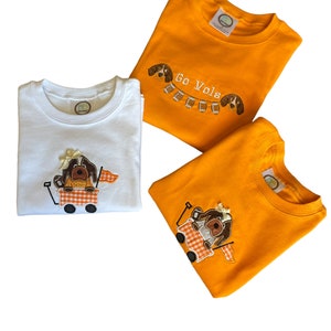 Puede incluir: Tres camisetas infantiles en blanco y naranja. La camiseta blanca tiene un perro bordado en un vag&oacute;n. Las camisetas naranjas tienen perros bordados y el texto "Go Vols". Las camisetas est&aacute;n dobladas y listas para usar.