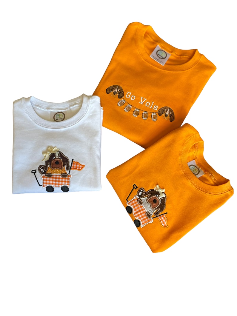 Puede incluir: Tres camisetas infantiles en blanco y naranja. La camiseta blanca tiene un perro bordado en un vag&oacute;n. Las camisetas naranjas tienen perros bordados y el texto "Go Vols". Las camisetas est&aacute;n dobladas y listas para usar.