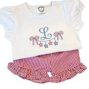 May include: White short-sleeved shirt with a blue and red embroidered design of stars and bows with the letter L in the centre. The shirt is paired with red and white gingham shorts with a ruffled hem.