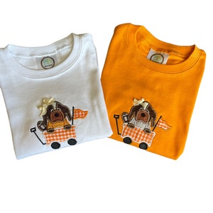 Puede incluir: Dos camisetas de manga corta, una blanca y otra naranja, cada una con un dise&ntilde;o bordado. El dise&ntilde;o representa un perro marr&oacute;n en un vag&oacute;n con un patr&oacute;n de cuadros, sosteniendo una bandera. Ambas camisetas tienen cuello redondo.
