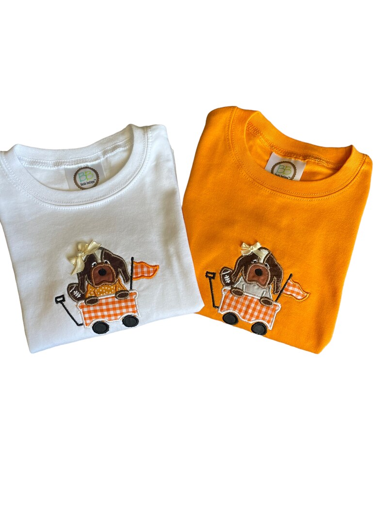 Puede incluir: Dos camisetas de manga corta, una blanca y otra naranja, cada una con un dise&ntilde;o bordado. El dise&ntilde;o representa un perro marr&oacute;n en un vag&oacute;n con un patr&oacute;n de cuadros, sosteniendo una bandera. Ambas camisetas tienen cuello redondo.