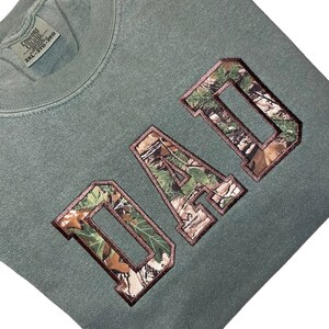 May include: A green sweatshirt with the word "DAD" in large, brown letters with a camouflage pattern inside the letters.