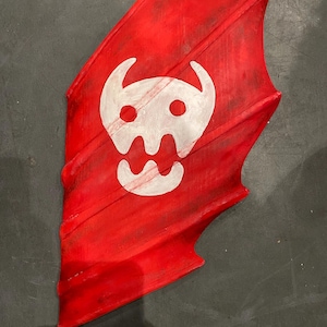 May include: A red, wing-shaped object featuring a white graphic of a horned, toothy creature face. The surface has a textured, slightly weathered appearance with visible ridges running across the wing structure, creating a dimensional effect.