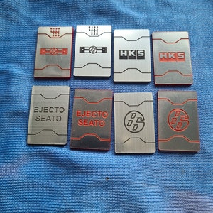 May include: Eight silver and red metal keychains with different designs. The designs include a gear shift pattern, the words "HKS", and a stylized logo.