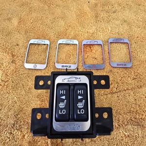 Puede incluir: Un panel de control negro del calentador del asiento del coche con molduras plateadas y cuatro piezas de molduras plateadas. El panel de control tiene dos juegos de botones "HI" y "LO" con un gráfico de coche. Las piezas de moldura plateadas tienen la palabra "BRZ" y un gráfico de coche.