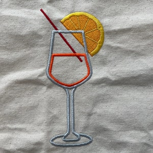 May include: Embroidered design of a cocktail glass with an orange slice and a red straw. The glass is outlined in gray with orange accents, and the orange slice is yellow. The design is on a beige fabric.