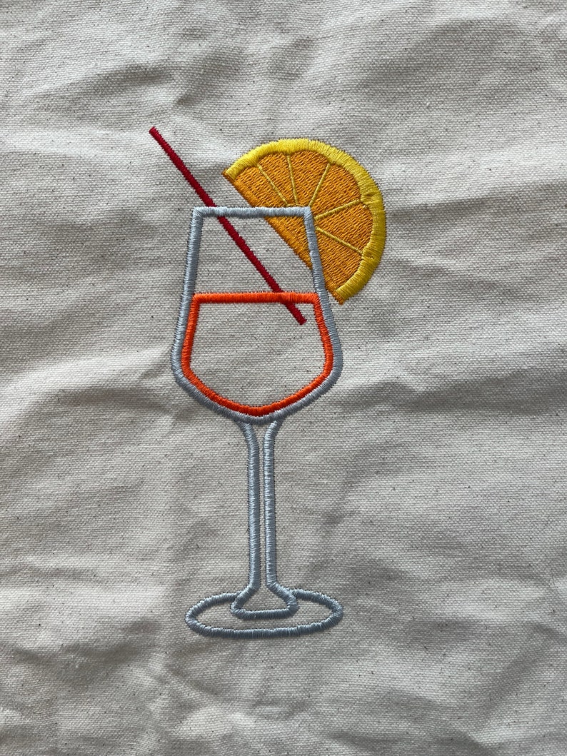 May include: Embroidered design of a cocktail glass with an orange slice and a red straw. The glass is outlined in gray with orange accents, and the orange slice is yellow. The design is on a beige fabric.