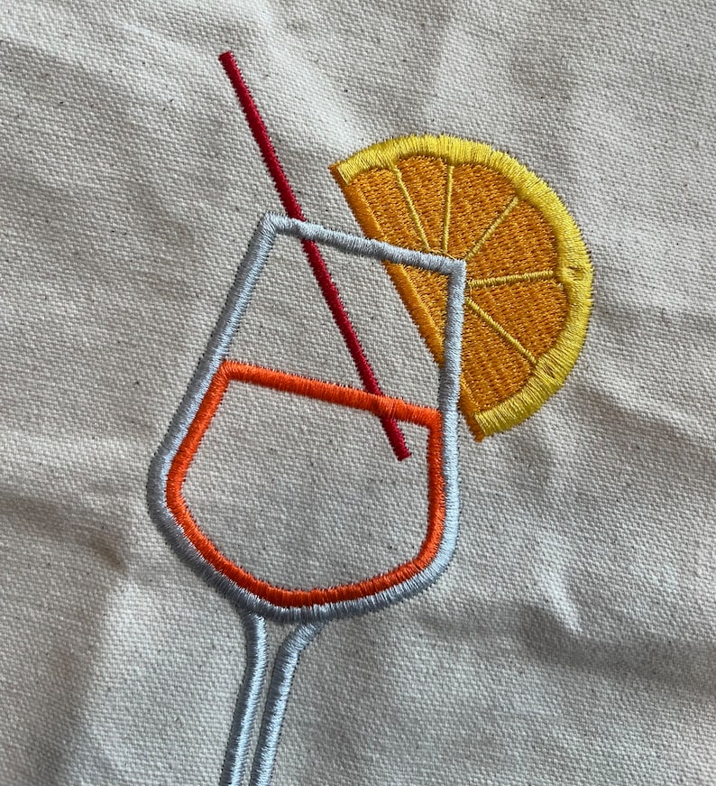 May include: Embroidered design of a cocktail glass with an orange slice and a red straw. The glass outline is silver with an orange interior. The orange slice is yellow and orange. The design is on a beige fabric.