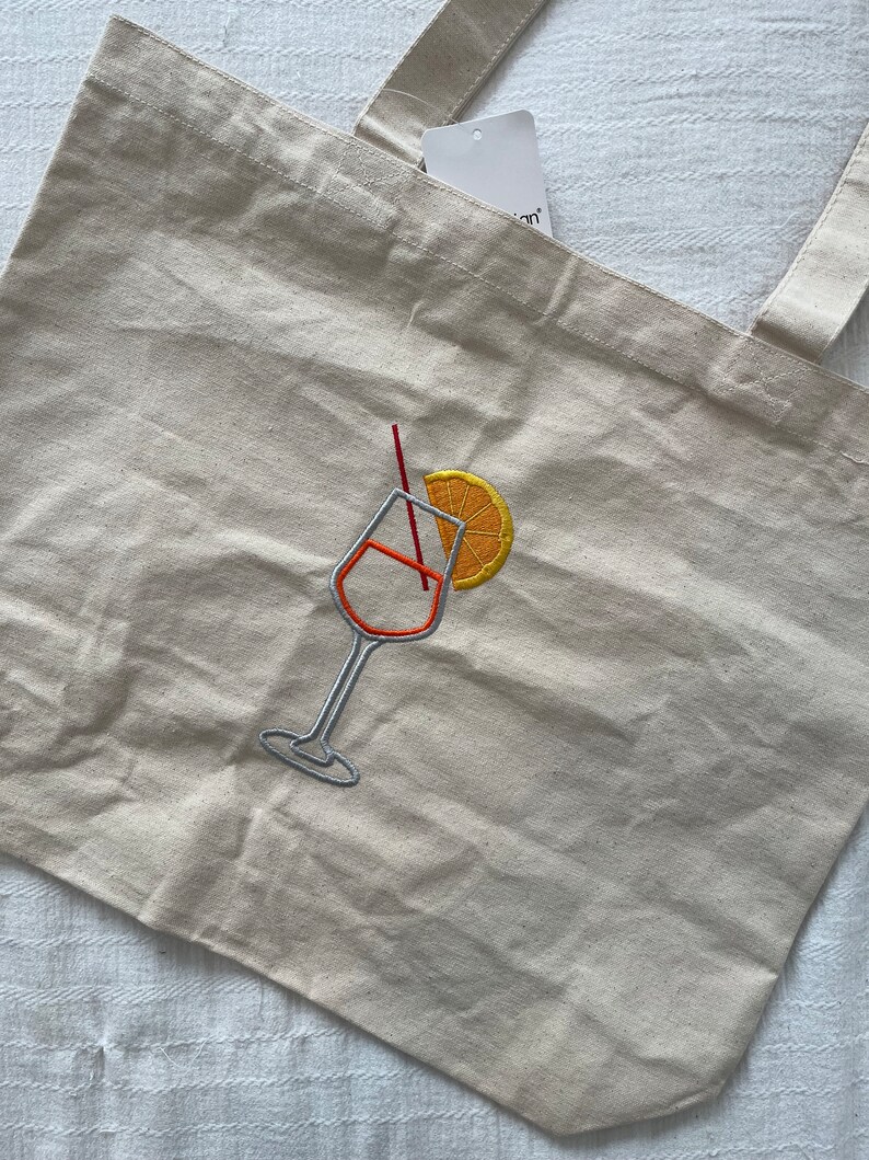 May include: Beige canvas tote bag with embroidered cocktail glass design. The design features an orange slice, a red straw, and a cocktail glass with orange and silver thread. The bag has two beige handles and a tag.