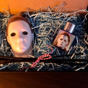 May include: A white mask with a human face and a black eye mask, a small glass bottle with a silver lid and a printed image of a man wearing a white mask, and black sticks in a blue and white shredded paper gift box.