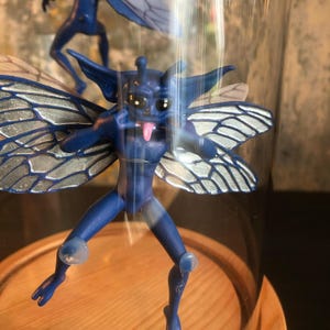 May include: A blue fairy figurine with detailed wings, inside a glass dome. The fairy has a playful expression, with its tongue sticking out. The figurine is set on a wooden base.