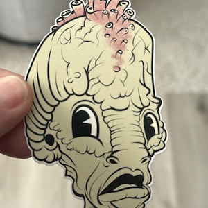 May include: A sticker featuring a cartoon illustration of a creature with a large head, wide eyes, and an open mouth. The creature has a light beige complexion with a cluster of pink and white tube-like growths on its head.