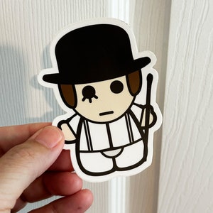 May include: A sticker featuring a cartoon character in a black bowler hat, white outfit with suspenders, and a cane. The character has brown hair and a single eye with a stylized design. The sticker is held by a hand.