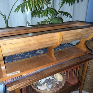 May include: A wooden planter box with a curved design and two shelves. The planter is made of light brown wood with a darker brown wood trim. The planter is sitting on a table with a woven basket and other items.