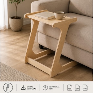 Op de afbeelding: Een lichtgekleurde houten C-vormige bijzettafel met een plat rechthoekig bovenoppervlak. De tafel heeft een Z-vormige steunstructuur die onder meubels schuift. Een beige keramische mok en een boek genaamd Kinfolk staan op de tafel.
