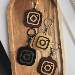 May include: Four keychains with different coloured Instagram logos. The logos are brown, gold, black, and brown with gold trim. The keychains are on a wooden surface.