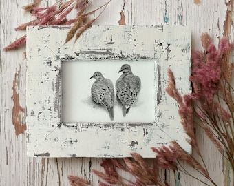 May include: A framed pencil drawing of two doves, set against a white background. The birds are depicted in detail, with visible feathers and shading. The frame is white with a distressed, rustic finish, adding a vintage aesthetic. Dried pink flowers are arranged around the frame.