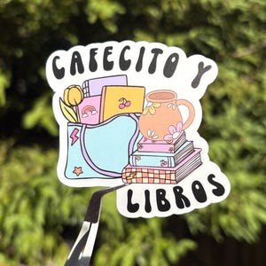 May include: A sticker with the words "Cafecito y Libros" in white, featuring a stack of books, a coffee mug, and a bag with a tulip, a rainbow, and a cherry. The sticker is held by tweezers against a green background.
