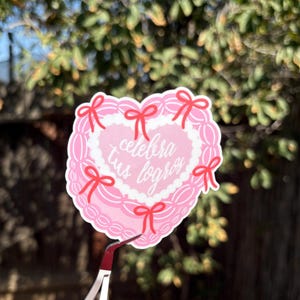 May include: A pink heart-shaped sticker with the Spanish text "celebra tus logros" in the center. The sticker has a white border and is decorated with red bows. The sticker is held by tweezers against a blurred green background.