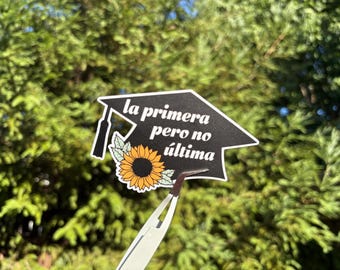 May include: A black graduation cap sticker featuring a yellow sunflower with green leaves and the white text "la primera pero no última" printed on the surface. The sticker is held by a pair of silver tweezers against a blurred green background.