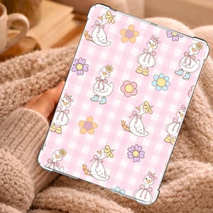 May include: A tablet case featuring a repeating pattern of white ducks wearing pink bows and yellow or blue rain boots, interspersed with colorful flowers on a light pink and white gingham background. The case has a clear, protective border.