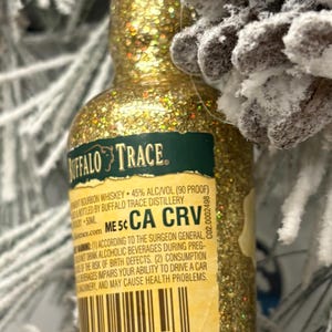 Puede incluir: Un adorno de botella de whisky bourbon Buffalo Trace en miniatura cubierto de purpurina dorada. La etiqueta incluye texto sobre el bourbon y advertencias. El adorno cuelga cerca de una pi&ntilde;a escarchada y ramas de hoja perenne.