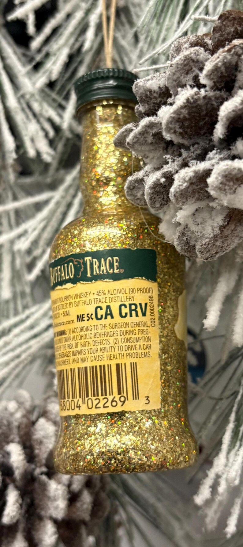 Puede incluir: Un adorno de botella de whisky bourbon Buffalo Trace en miniatura cubierto de purpurina dorada. La etiqueta incluye texto sobre el bourbon y advertencias. El adorno cuelga cerca de una pi&ntilde;a escarchada y ramas de hoja perenne.