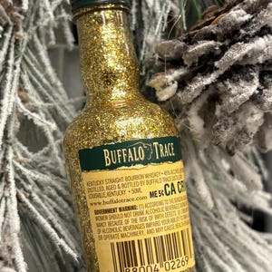Puede incluir: Un adorno de botella de whisky bourbon Buffalo Trace en miniatura, cubierto de purpurina dorada. La etiqueta es verde y blanca, con el nombre de la marca y la informaci&oacute;n del producto. La botella cuelga de una cuerda de hilo, sobre una rama de pino nevada.