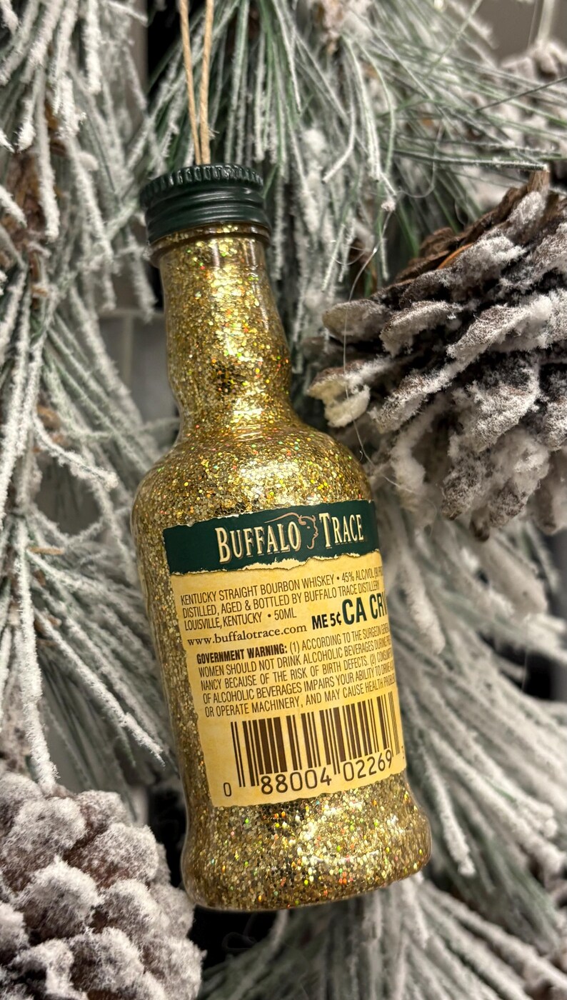 Puede incluir: Un adorno de botella de whisky bourbon Buffalo Trace en miniatura, cubierto de purpurina dorada. La etiqueta es verde y blanca, con el nombre de la marca y la informaci&oacute;n del producto. La botella cuelga de una cuerda de hilo, sobre una rama de pino nevada.