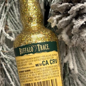 Puede incluir: Una botella en miniatura de whisky bourbon Buffalo Trace, cubierta de purpurina dorada. La etiqueta es visible, con texto como "Buffalo Trace" y "Kentucky Straight Bourbon Whiskey". La botella es de 50 ml.