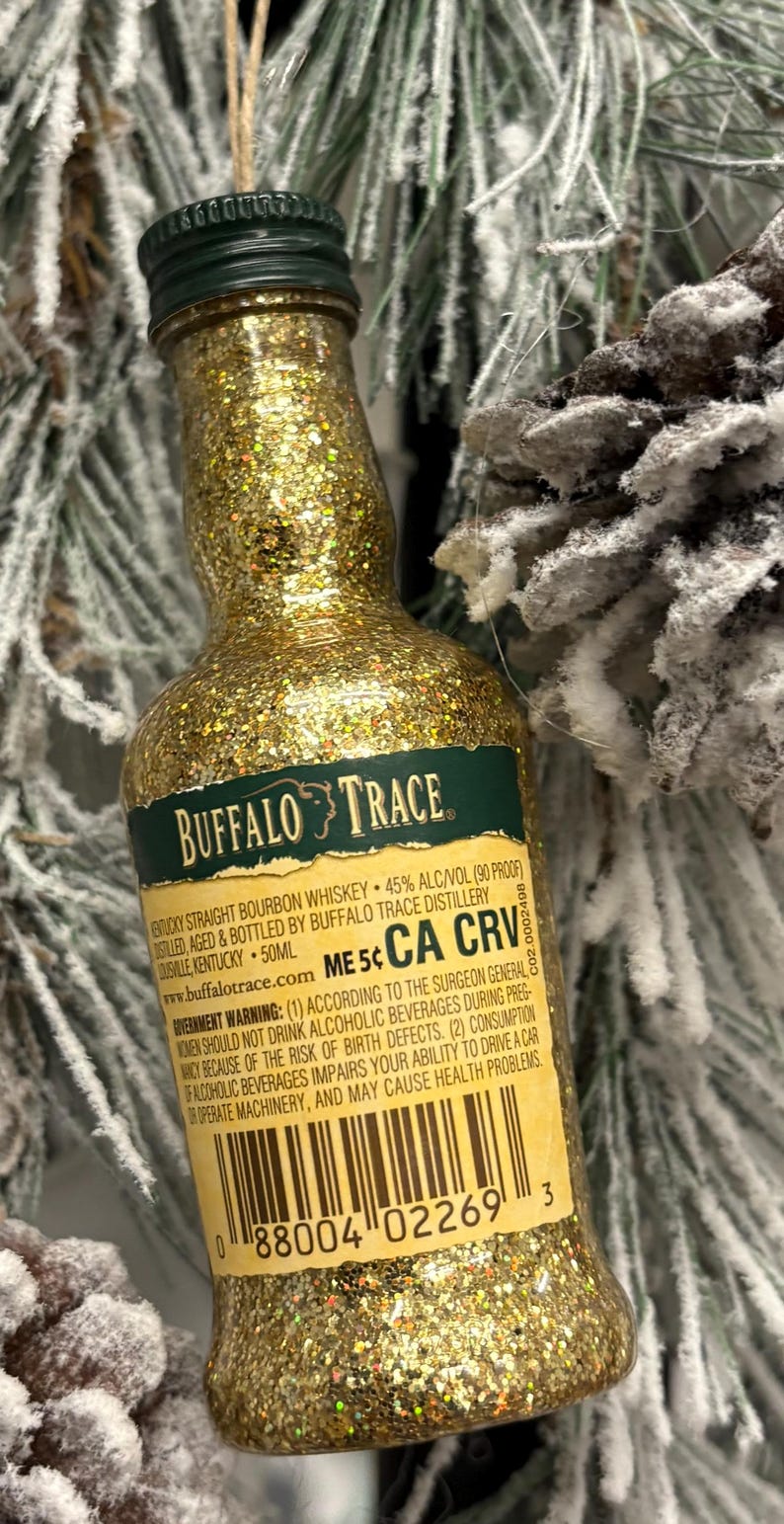 Puede incluir: Una botella en miniatura de whisky bourbon Buffalo Trace, cubierta de purpurina dorada. La etiqueta es visible, con texto como "Buffalo Trace" y "Kentucky Straight Bourbon Whiskey". La botella es de 50 ml.
