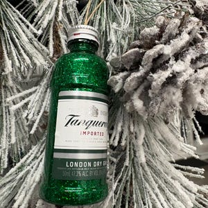 Puede incluir: Adorno navide&ntilde;o en forma de botella de ginebra en miniatura, cubierta de purpurina verde. La etiqueta dice "Tanqueray Imported London Dry Gin", con un volumen de 50 ml. El adorno cuelga de una cuerda, sobre un fondo de pino helado y una pi&ntilde;a.